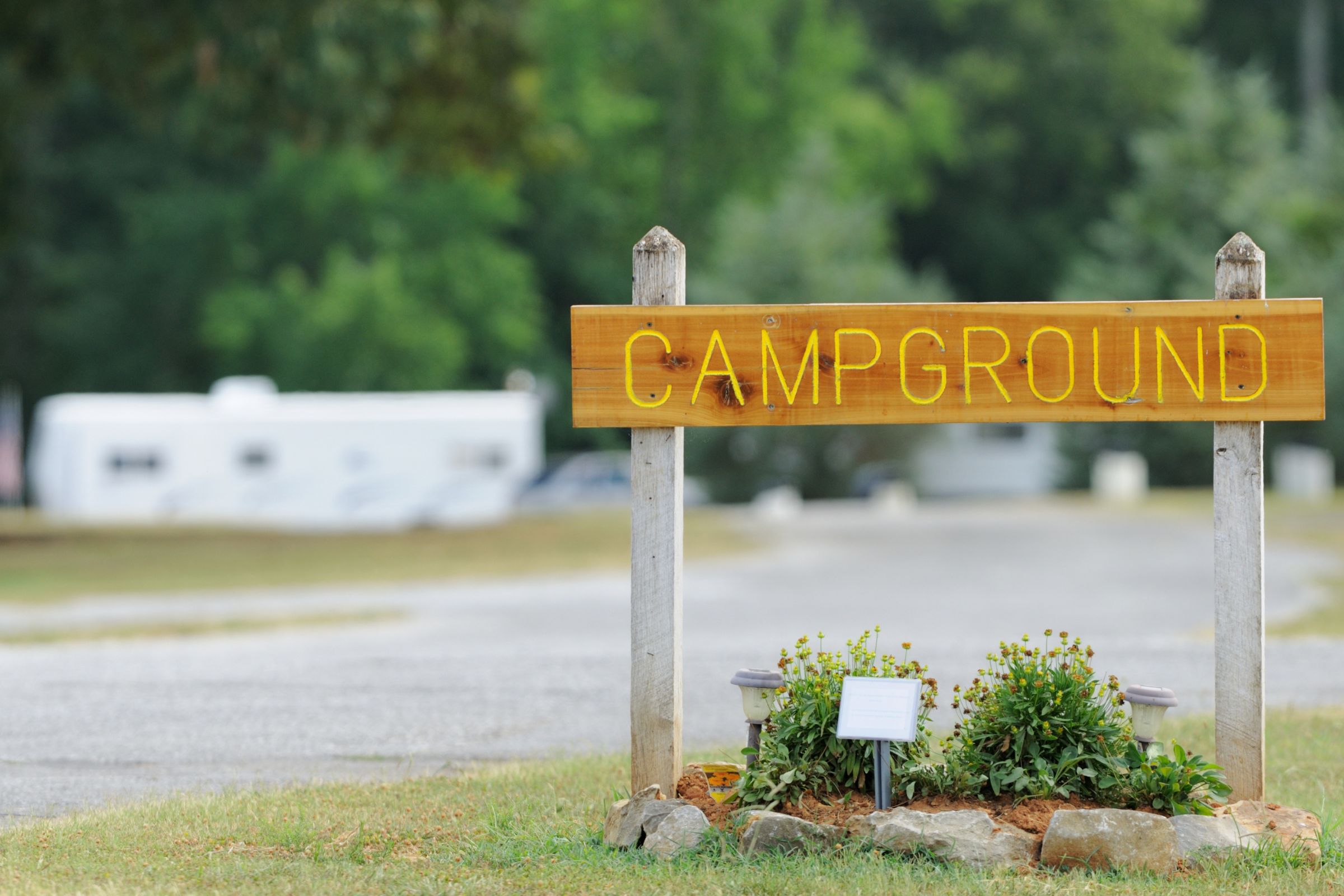 Explore Gear | Wooden campground sign with yellow letters, surrounded by greenery, and RVs visible in the background.