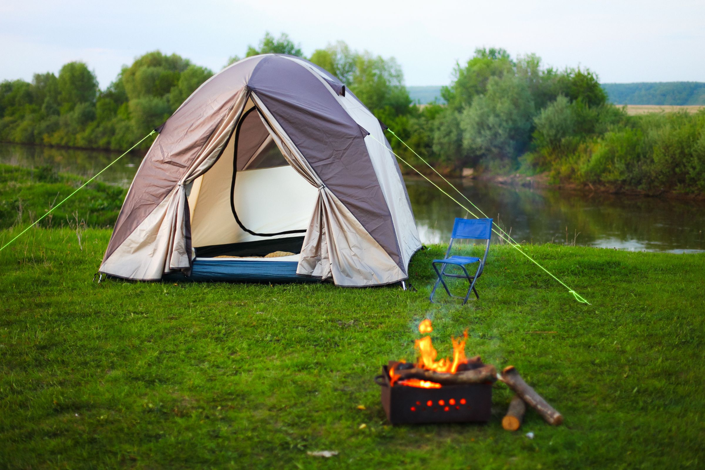 ExploreGear.com | Camping setup with a pitched tent, a blue chair, and a small campfire near a scenic riverside surrounded by greenery.