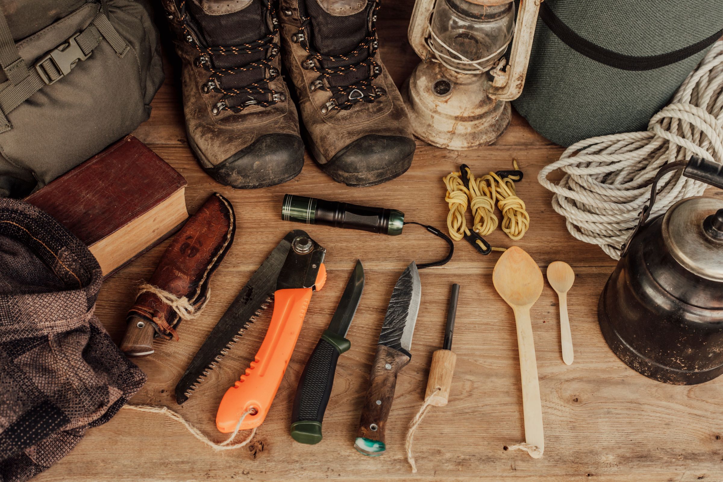 ExploreGear.com | Flat lay of essential camping tools and gear, including hiking boots, ropes, knives, a lantern, and a saw, arranged on a wooden surface.