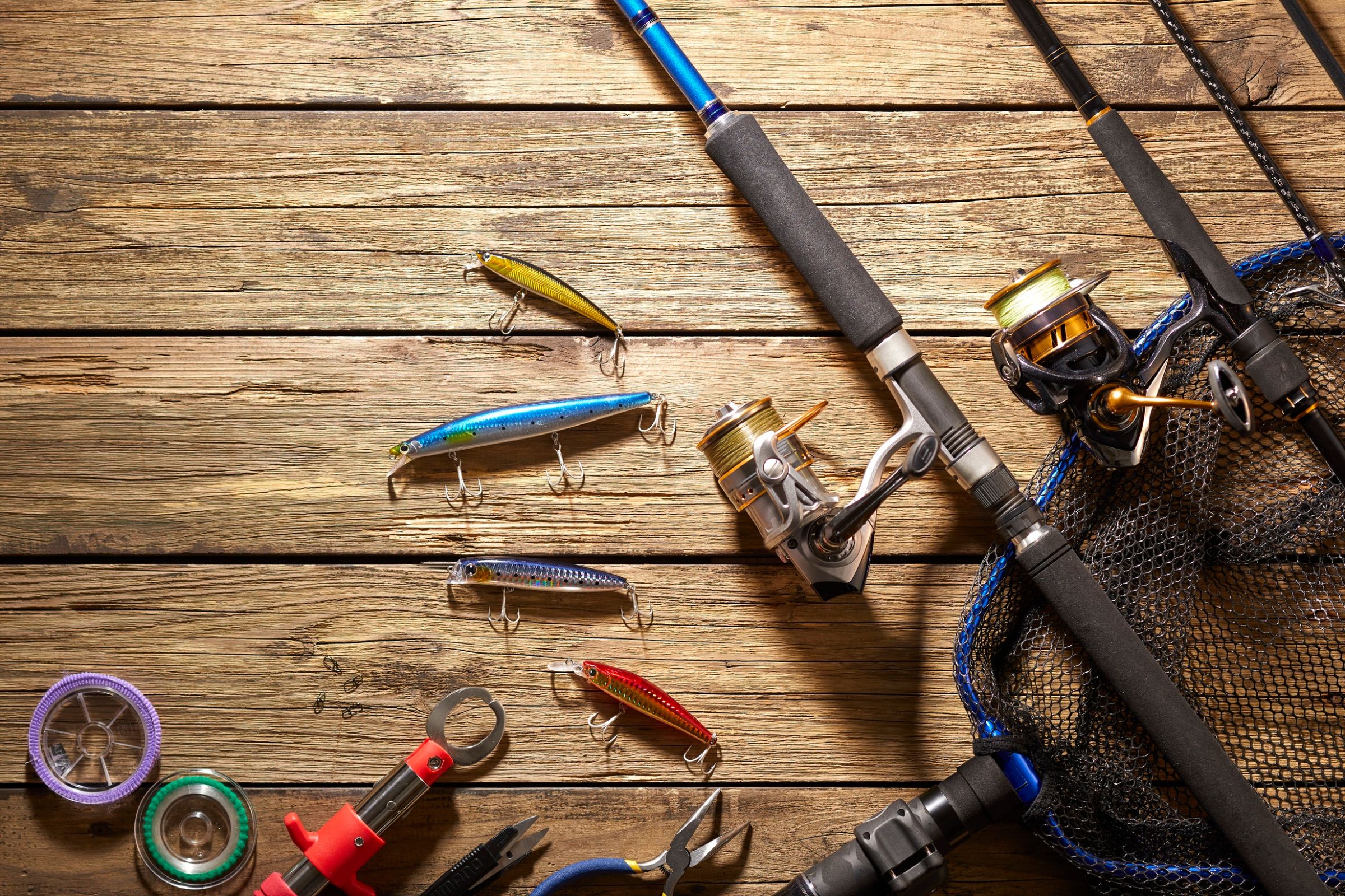 A collection of fishing gear on a wooden surface, including spinning rods, colorful lures, a fishing net, pliers, a fish grip, and spools of fishing line, showcasing essential tools for anglers.