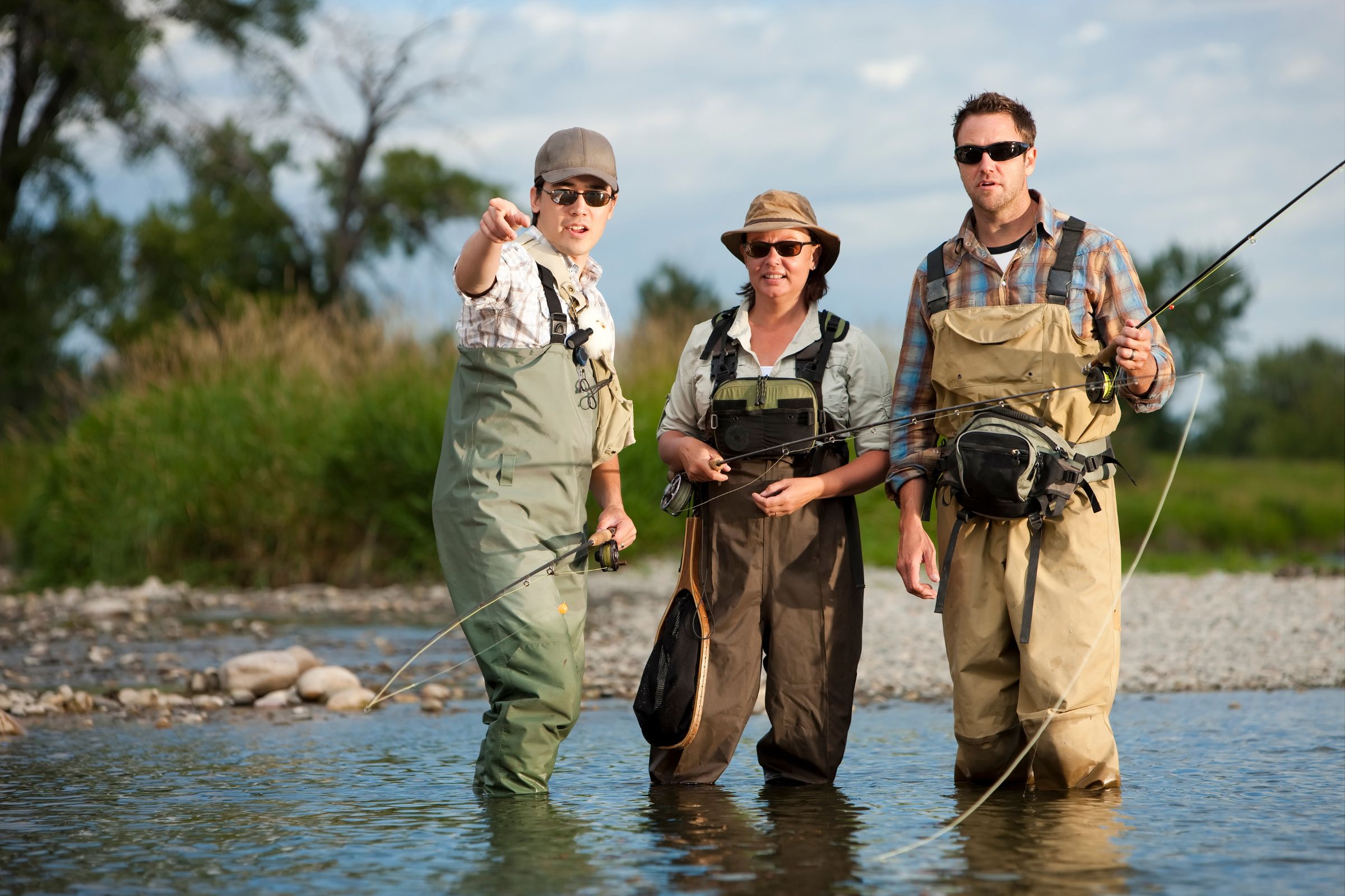 A fishing guide standing in shallow water with two clients, all wearing waders and holding fishing rods, as the guide gestures and explains techniques amidst a scenic riverside backdrop.