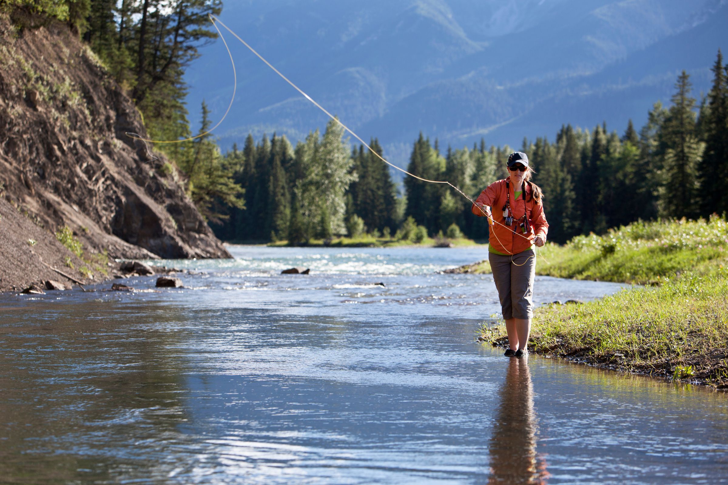 A woman fly fishing in a clear mountain stream, surrounded by lush green trees and distant forested hills under a bright blue sky.