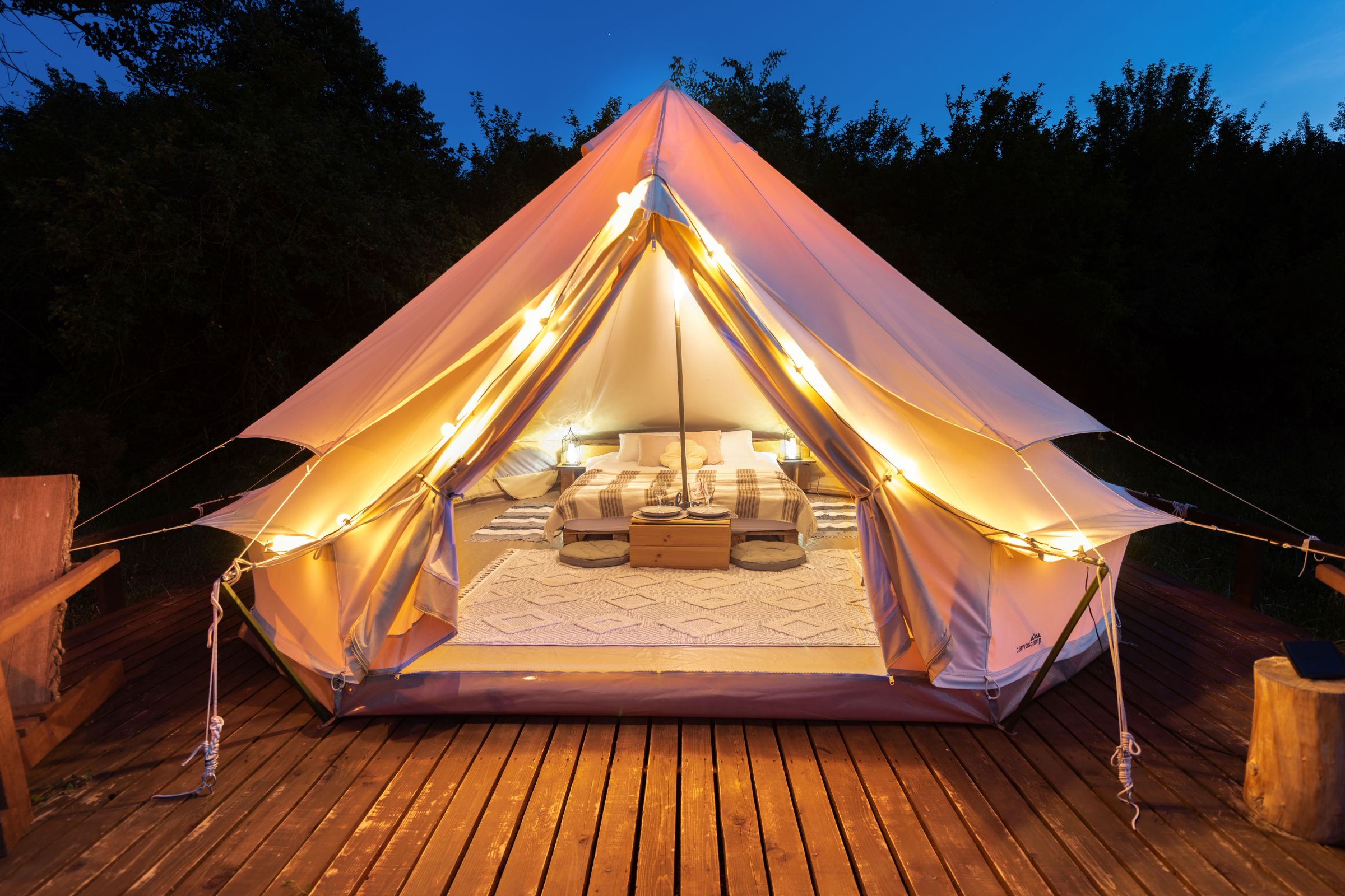 Cozy glamping tent illuminated by string lights, featuring a comfortable bed and stylish interior decor on a wooden deck surrounded by nature.