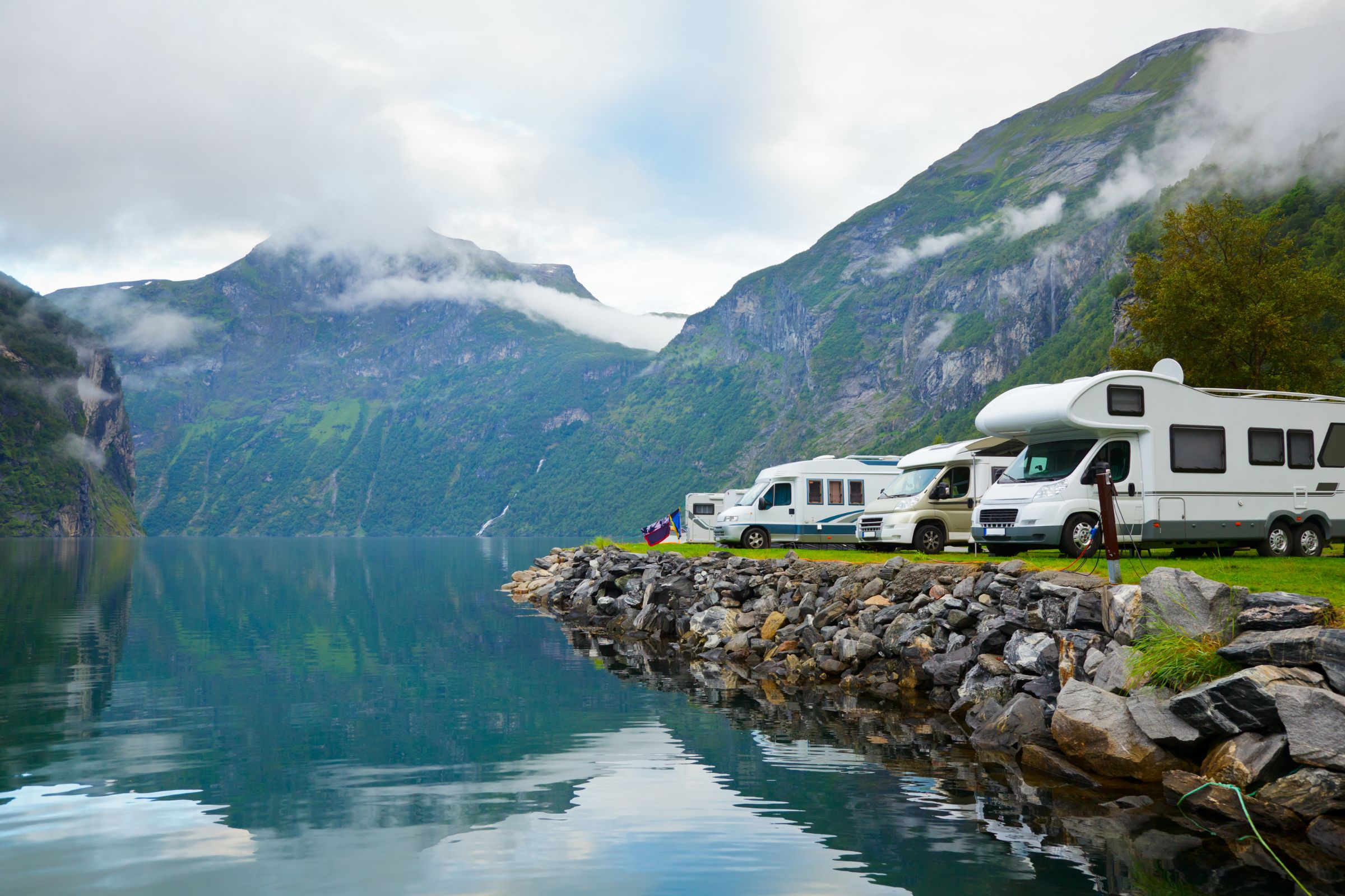 ExploreGear.com | RVs parked along a scenic lakeside with rocky edges, surrounded by lush green mountains and misty skies.