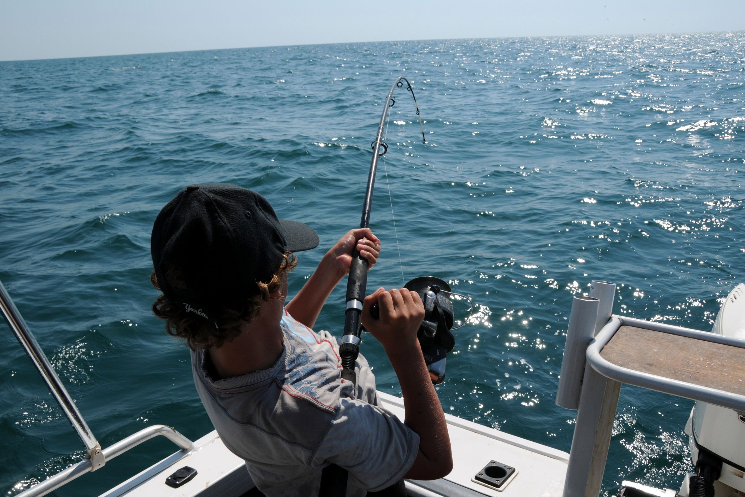 A young angler wearing a black cap reels in a fishing rod on a boat, surrounded by the vast, sparkling blue ocean under a clear sky.