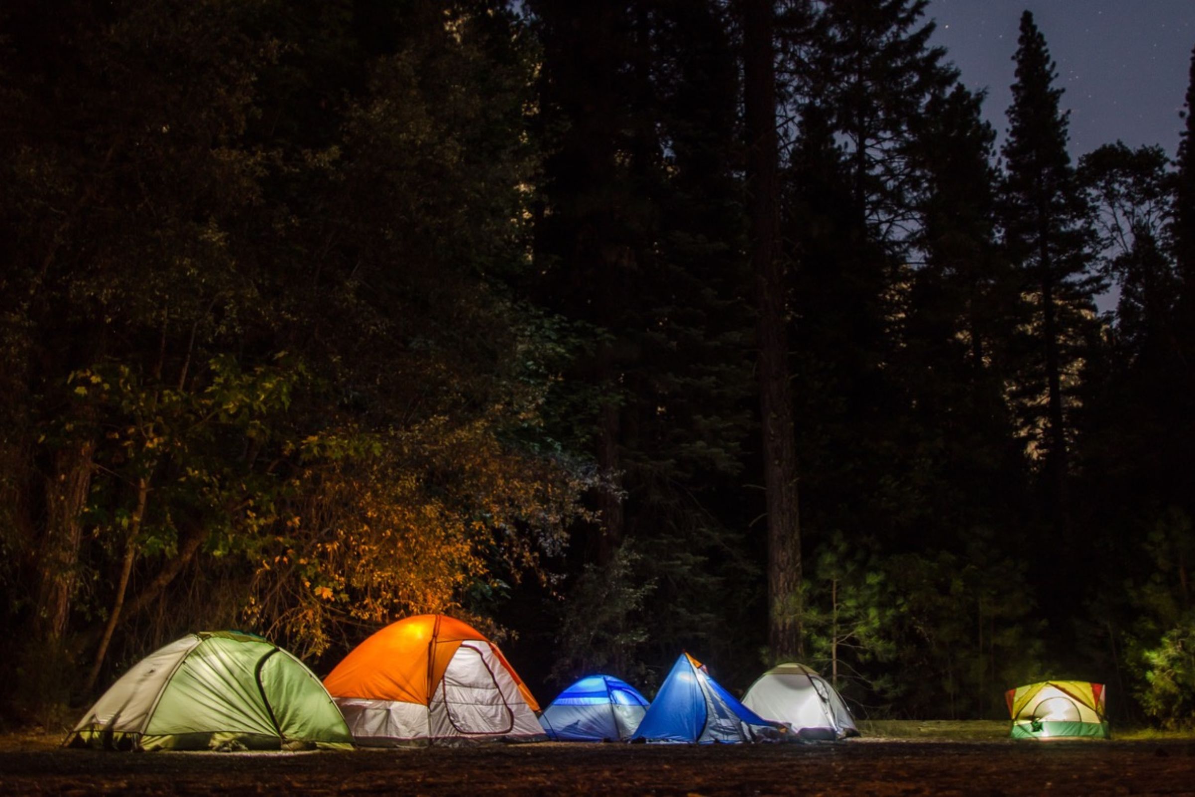 ExploreGear.com | A group of colorful tents illuminated at night in a forested campsite under a starry sky.