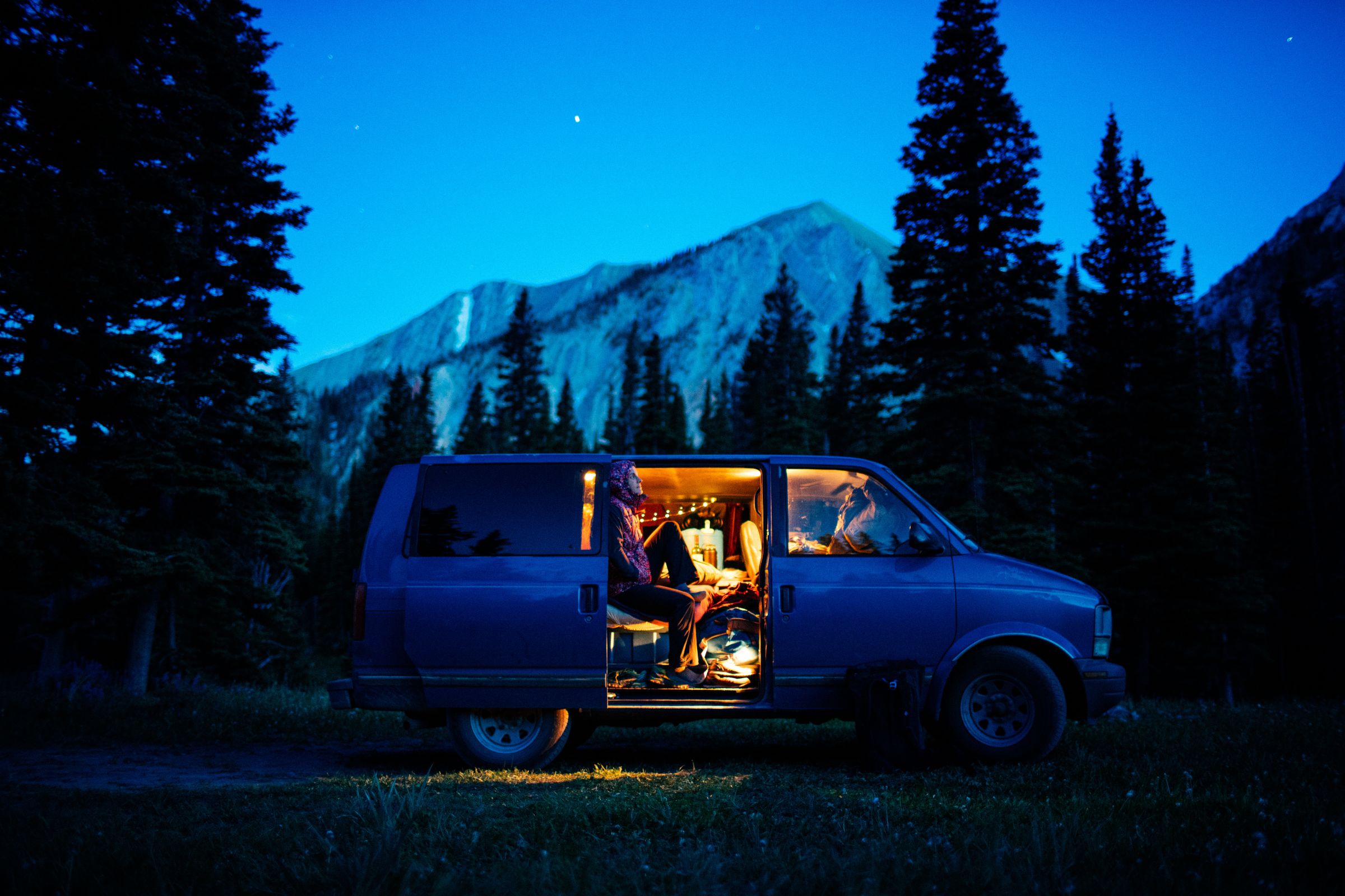 ExploreGear.com \ A lit camper van parked in a forest at night, surrounded by tall trees and mountains under a starry sky.